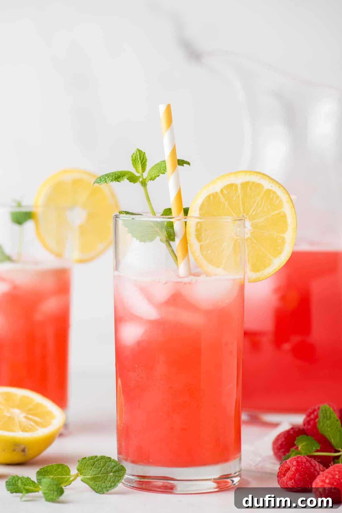 A glass of vibrant pink raspberry lemonade, garnished with a fresh lemon slice, a sprig of mint, and a striped paper straw.