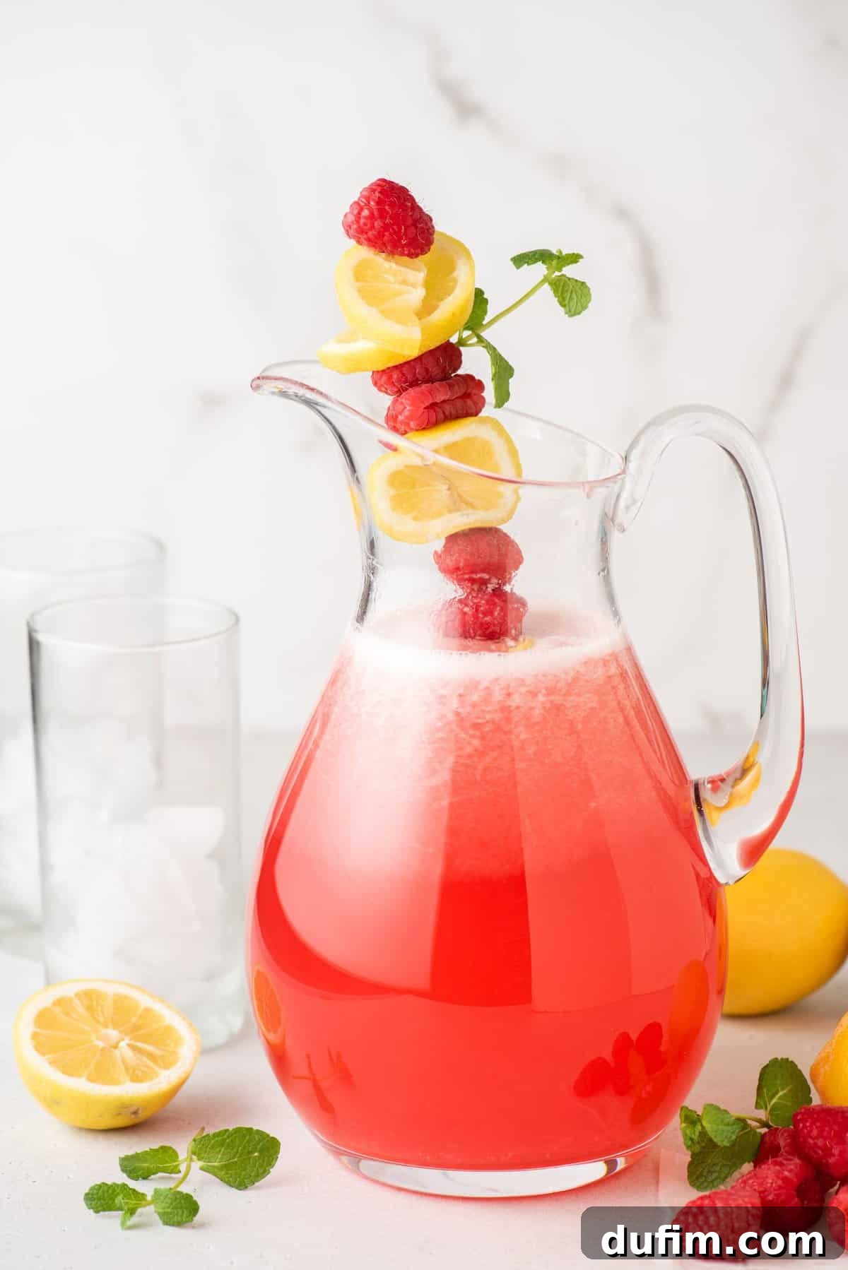 A large glass pitcher of homemade raspberry lemonade, beautifully garnished with a skewer of fresh raspberries, vibrant lemon slices, and a fragrant sprig of mint, ready for serving.