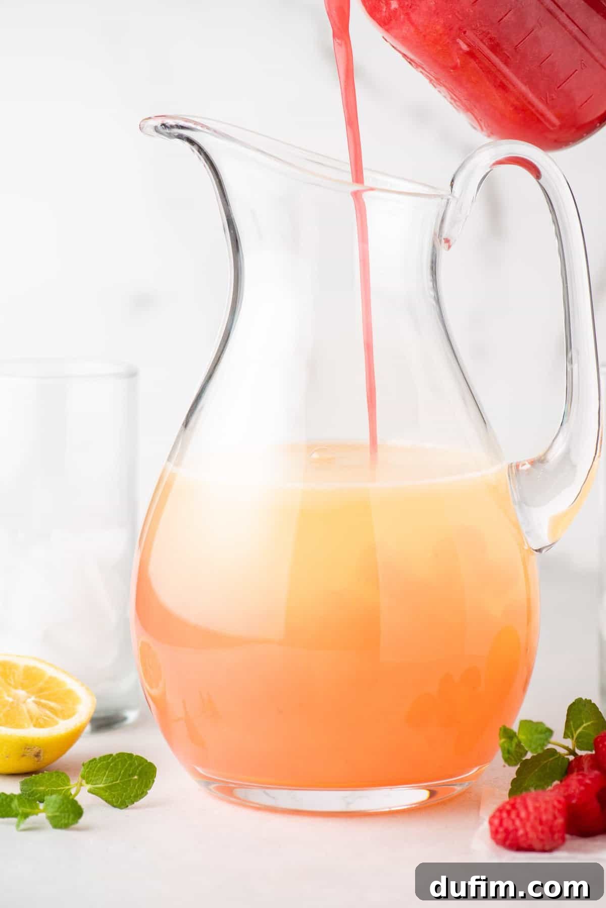 A stream of rich pink raspberry juice is being poured into a large glass pitcher already containing clear lemon juice, as the final step in combining the lemonade ingredients.