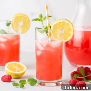 A glass of homemade raspberry lemonade with ice, garnished with a paper straw, a sprig of mint, and a lemon slice.