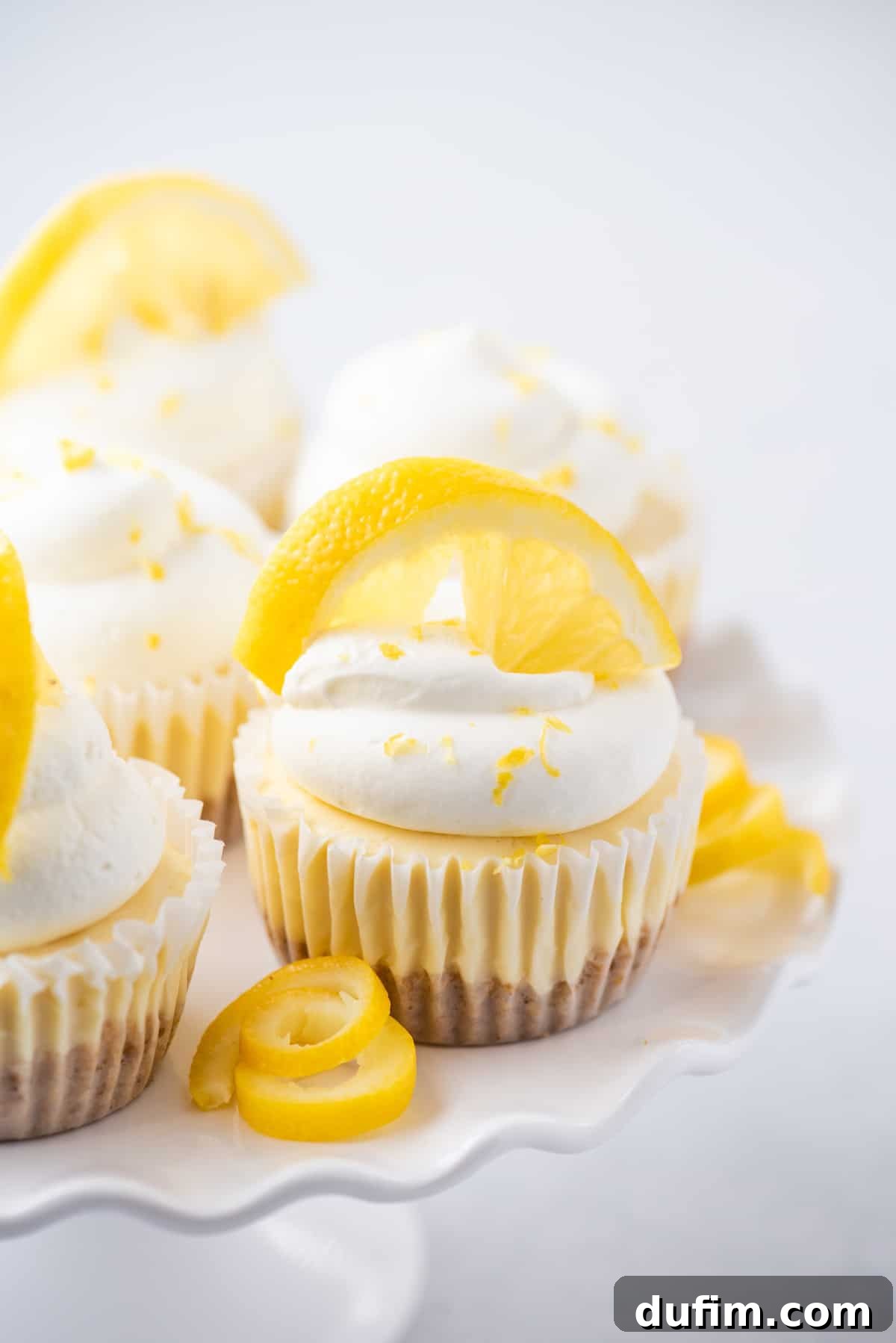 Zesty Lemon Cheesecake Bites 7 Mini lemon cheesecakes topped with whipped cream and lemon zest arranged on white cake stand