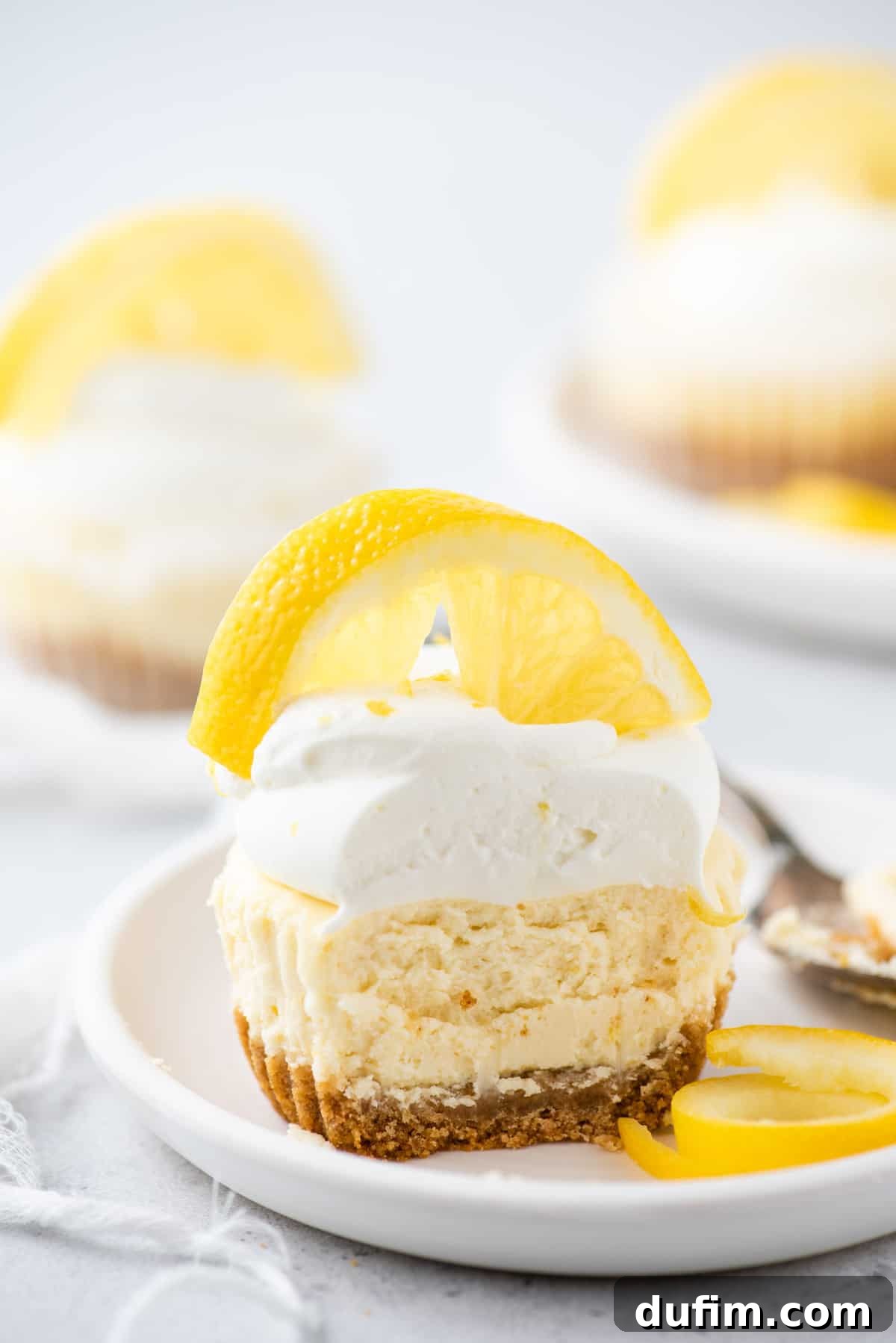 Zesty Lemon Cheesecake Bites 8 Mini lemon cheesecake topped with whipped cream and lemon slice with bite removed on white plate