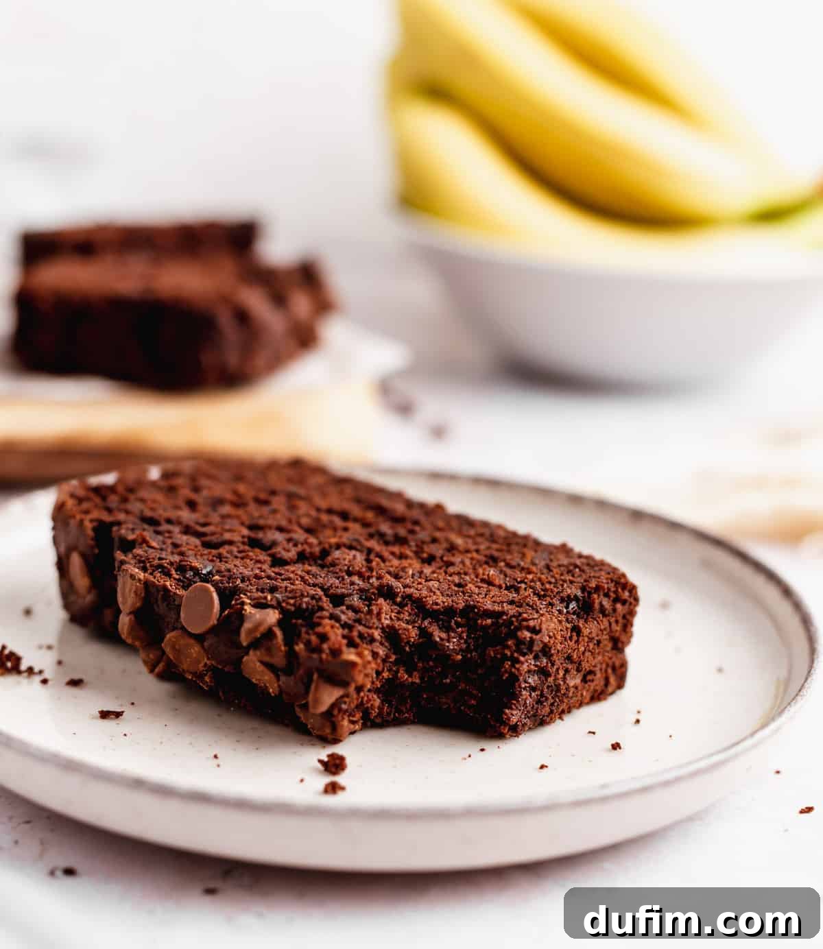 A close-up of a single slice of Double Chocolate Banana Bread on a white plate, with a bite taken out of the corner, emphasizing its moist interior and visible chocolate chips.