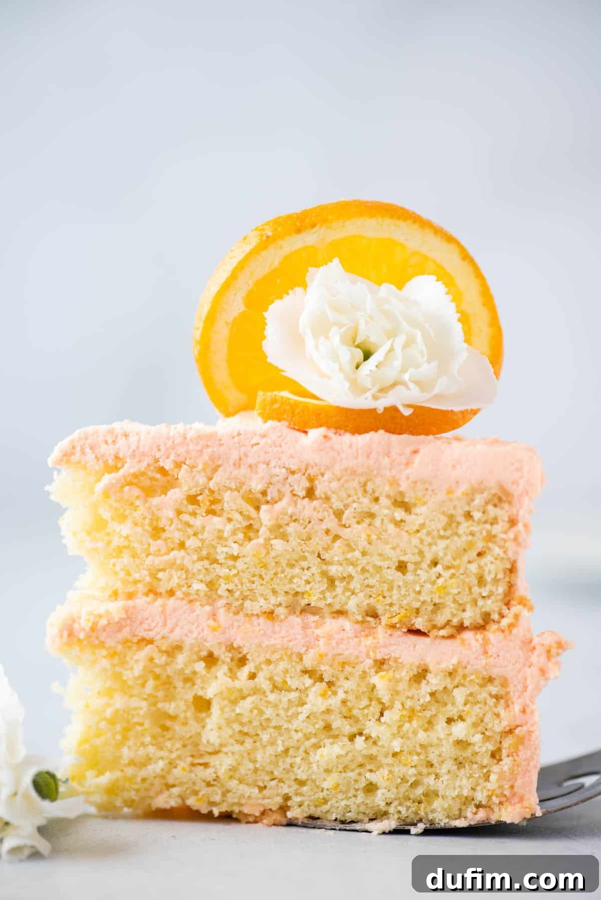 A close-up of a perfectly sliced orange cake, showing two moist layers separated by a generous helping of cream cheese frosting. The slice is artfully decorated with a fresh orange segment and a small white edible flower, presented on a sleek metal spatula.