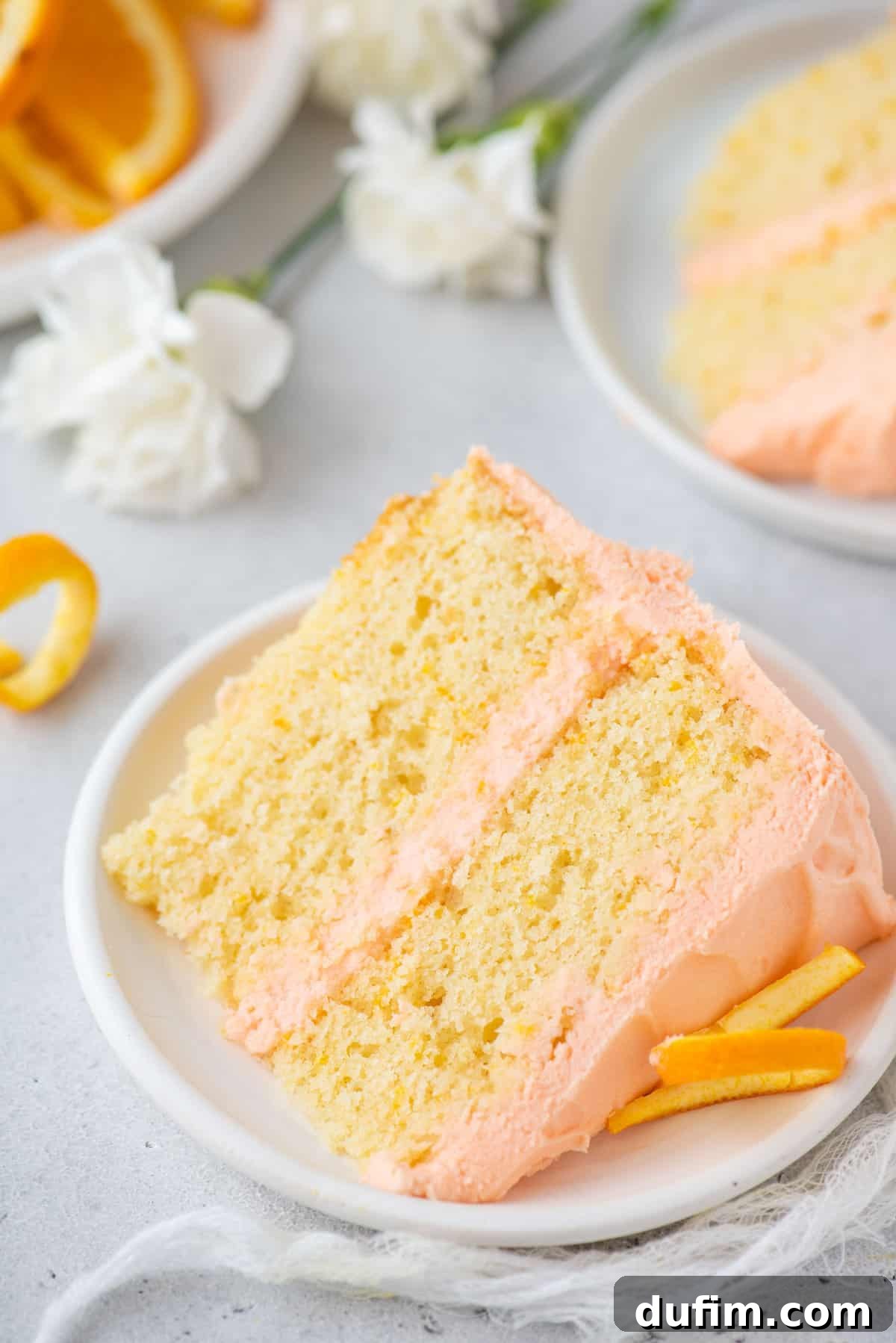 A generous slice of moist orange cake, featuring visible orange zest specks and a thick layer of creamy white frosting, laid on its side on a clean white plate. The soft texture and vibrant color make it look irresistible.