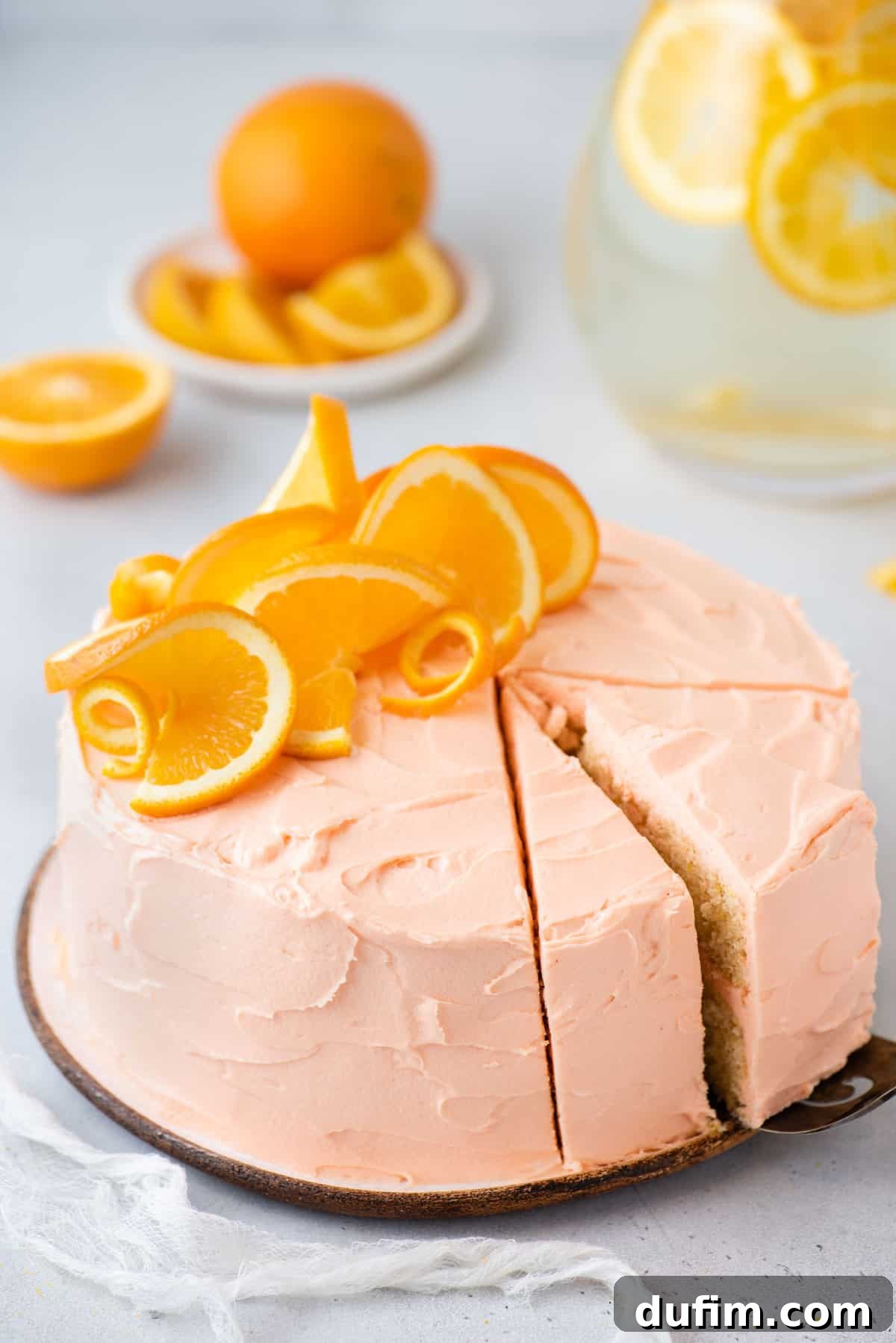A freshly baked, two-layer orange cake, beautifully frosted with cream cheese icing and decorated with vibrant orange slices and orange peel curls on top. It sits on a round white plate against a soft white background, ready to be served.