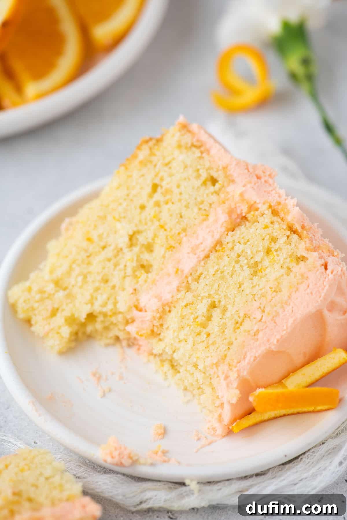 A delectable slice of orange cake, with a small bite removed, lies on its side on a simple white plate. The visible layers of cake and frosting, along with the fresh orange zest, highlight its inviting texture and bright flavor.