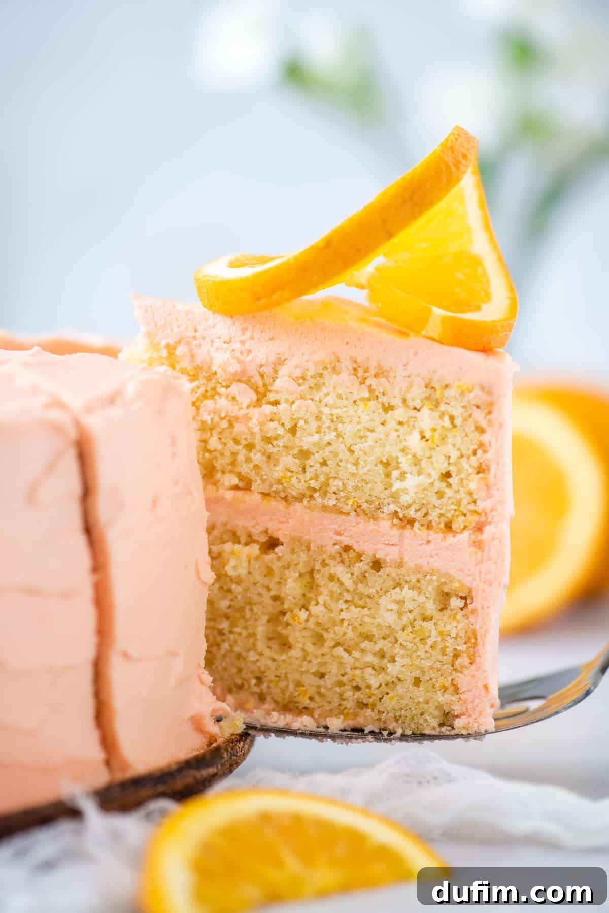 A single slice of orange cake sits on a metal spatula, ready for serving. It's elegantly decorated with a fresh orange slice and a small white flower, highlighting its delicate texture and vibrant citrus appeal. A full, frosted orange cake can be seen blurred in the background.