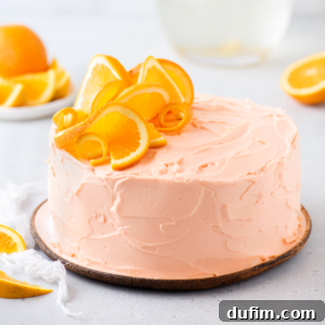 A round Orange Cake with smooth, light orange frosting is decorated on top with fresh orange slices and peel curls. The cake sits on a brown plate, with extra orange slices in the background for a vibrant display.