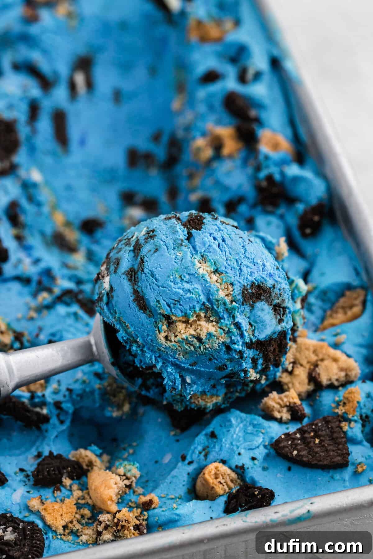 Scooping Homemade Cookie Monster Ice Cream from a pan, revealing its vibrant blue color and generous chunks of Oreos and chocolate chip cookies.