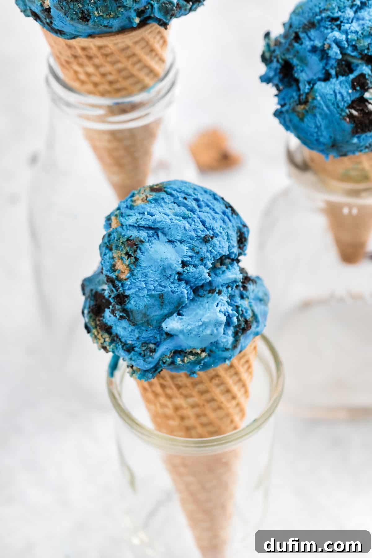 Three glass jars holding cones piled high with blue Cookie Monster Ice Cream, showcasing its inviting color and texture.