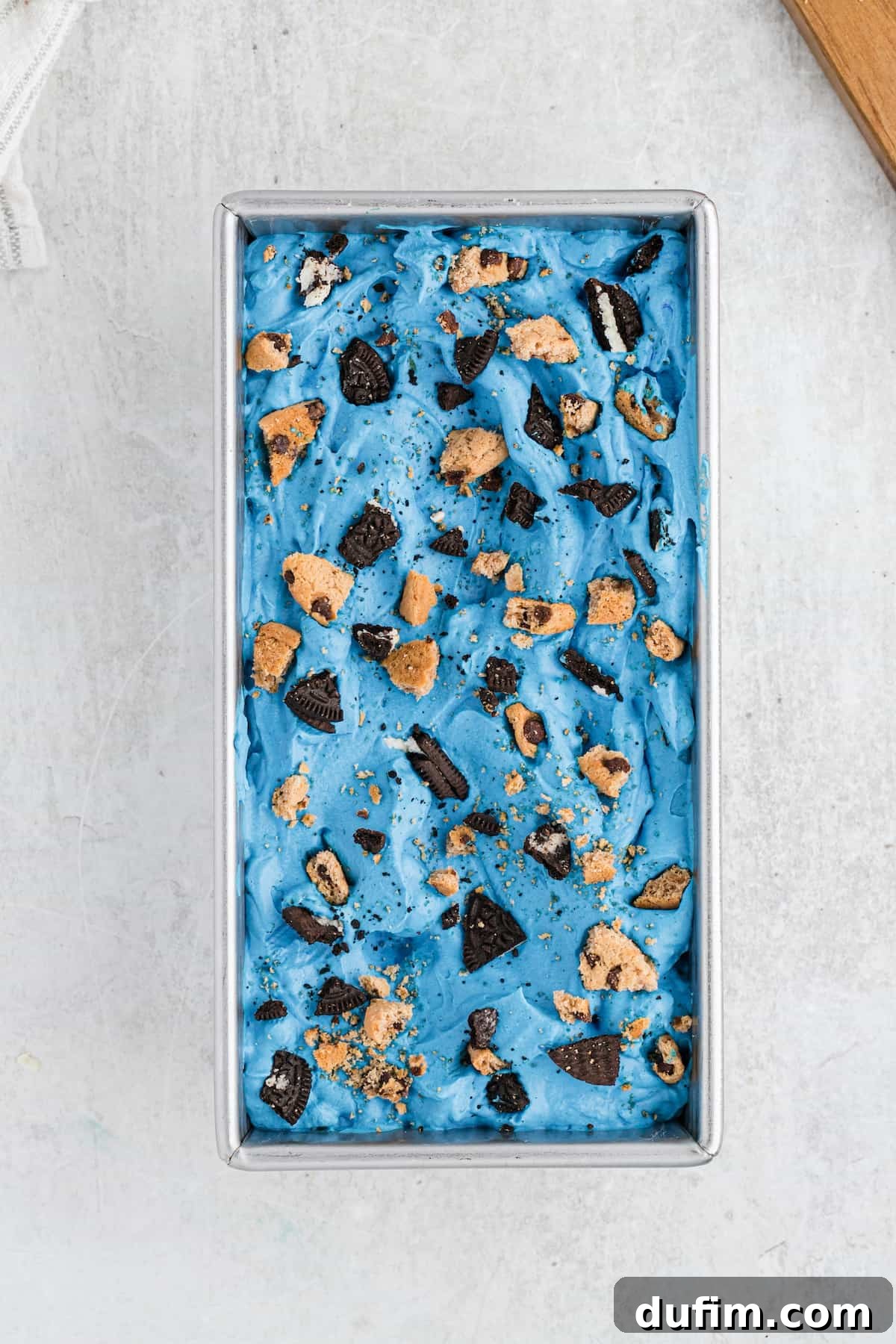 A loaf pan filled with beautifully frozen Cookie Monster Ice Cream, topped with extra cookie pieces, ready to be scooped.
