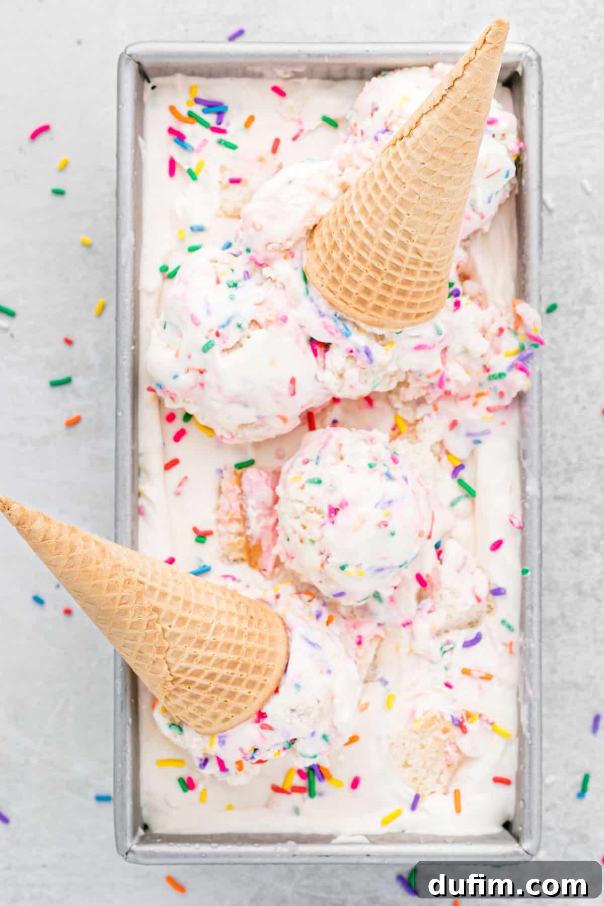 This no-churn Birthday Cake Ice Cream is loaded with colorful sprinkles and cupcakes! So easy to make, and you don't need an ice cream maker! A loaf pan filled with birthday cake ice cream, with scoops and ice cream cones arranged on top, ready to serve.