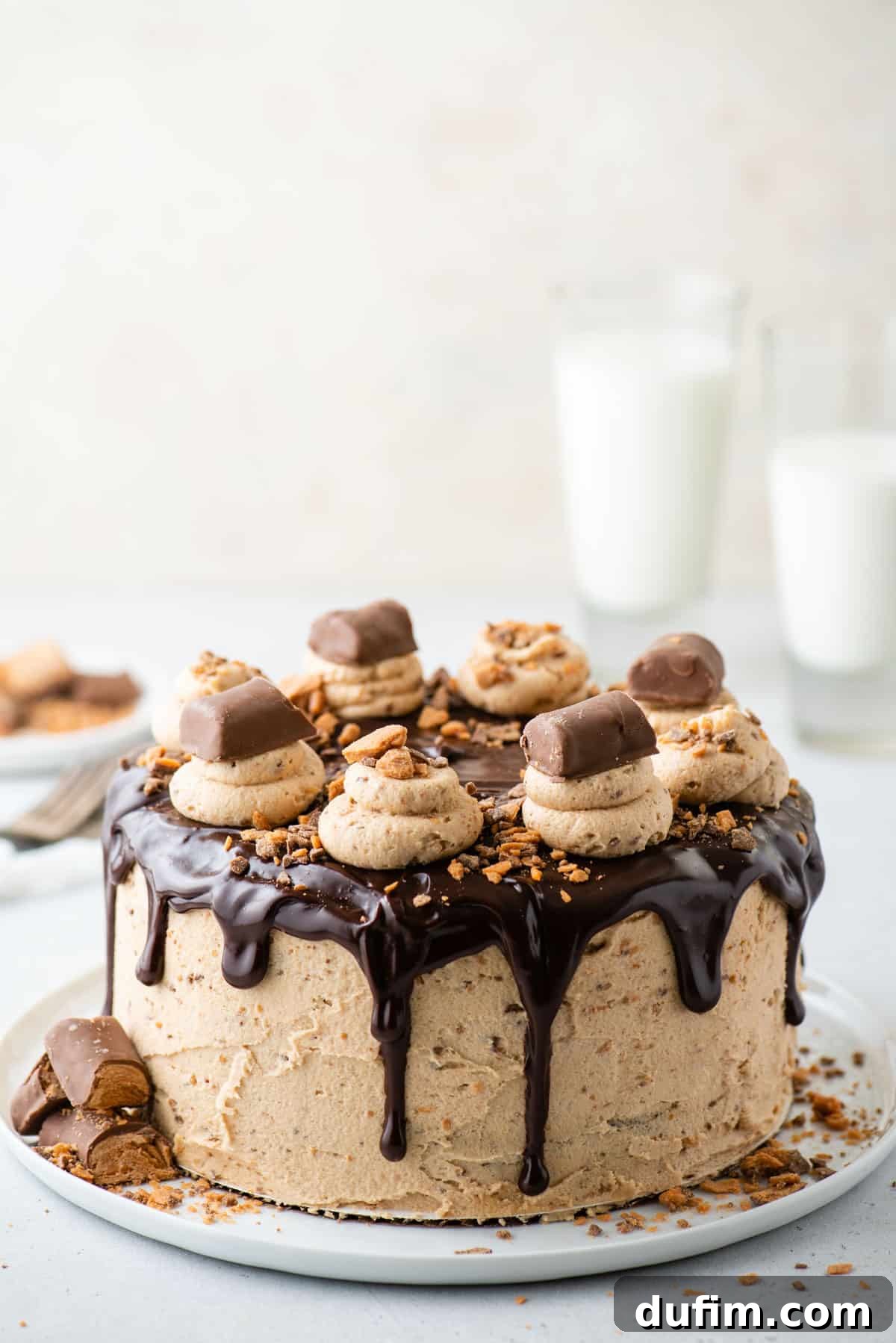 A stunning, multi-layered Butterfinger cake presented elegantly on a cake plate, with blurred glasses of milk in the background. The cake boasts a smooth frosting and a rich ganache drizzle, topped with crumbled Butterfinger pieces.