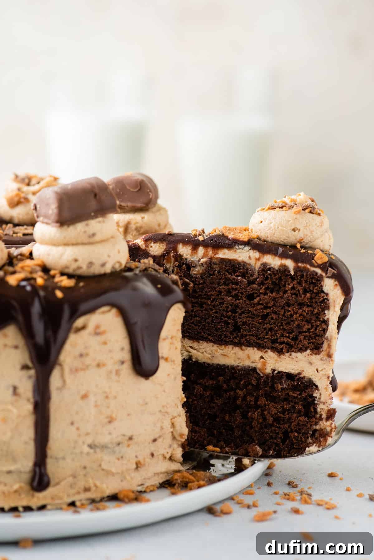 A hand gently lifts a perfect slice of the Butterfinger cake from the main dessert, highlighting the beautiful layers and generous topping.