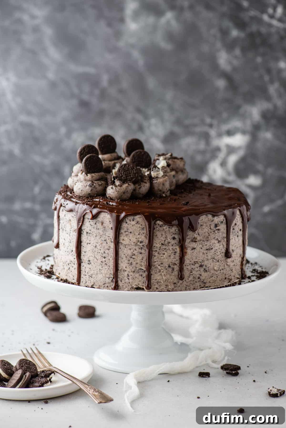 A beautifully decorated whole Oreo cake displayed on a classic white cake stand, ready to be served. It features rich chocolate layers, creamy Oreo frosting, and a glossy chocolate ganache drip.