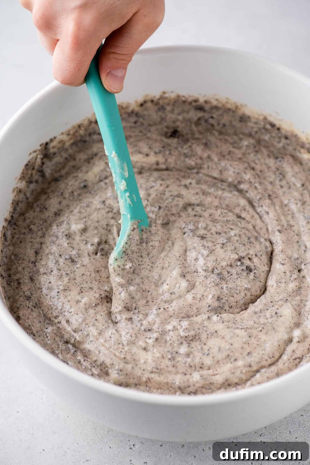 A close-up view of hands mixing rich Oreo cake batter in a large stainless steel bowl, highlighting the texture and cookie pieces.