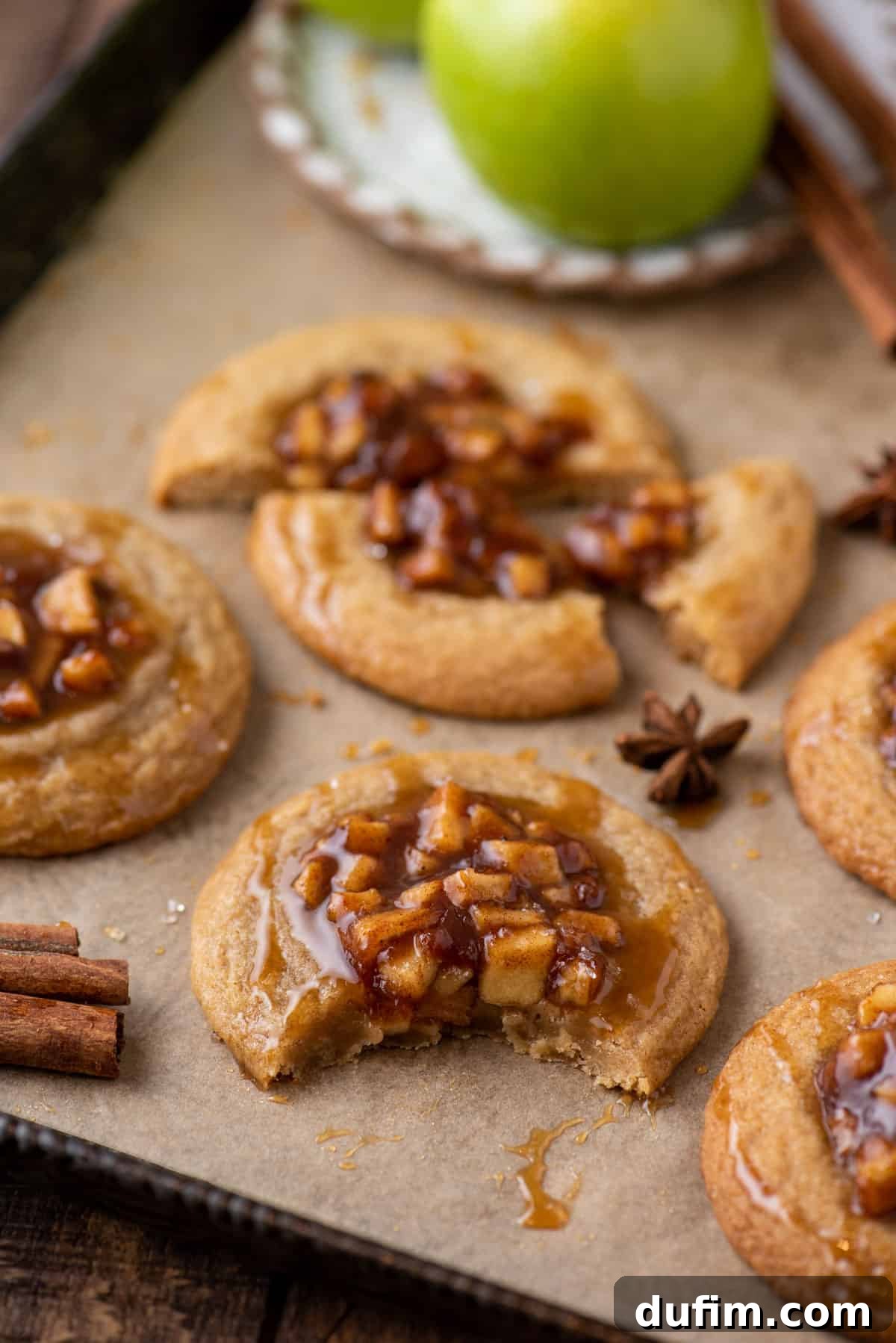 Freshly baked apple pie cookies arranged on a baking sheet, surrounded by fall spices like cinnamon sticks and star anise, evoking a warm autumnal ambiance.