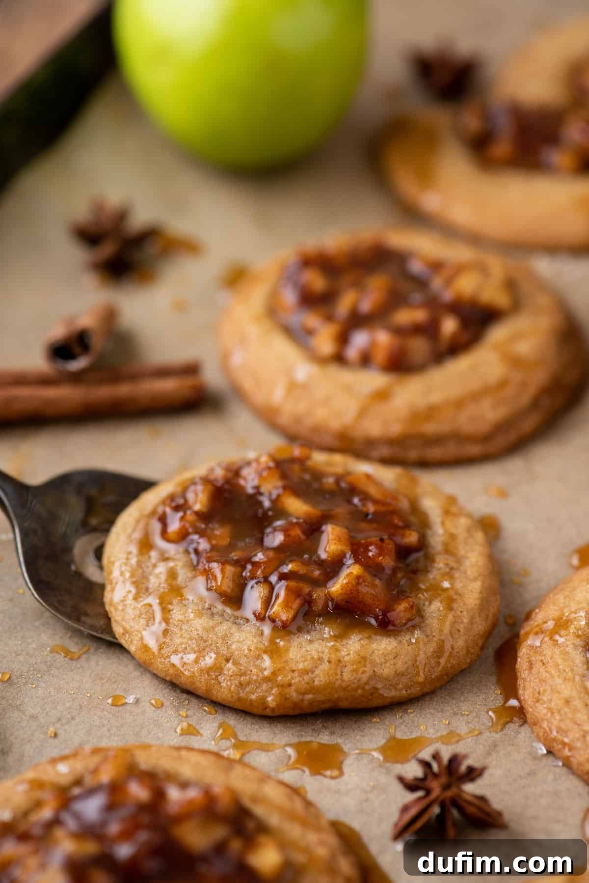 Close-up of baked apple pie cookies on a baking sheet, with one cookie being gently lifted by a spatula, showcasing its golden-brown edges and luscious apple filling.