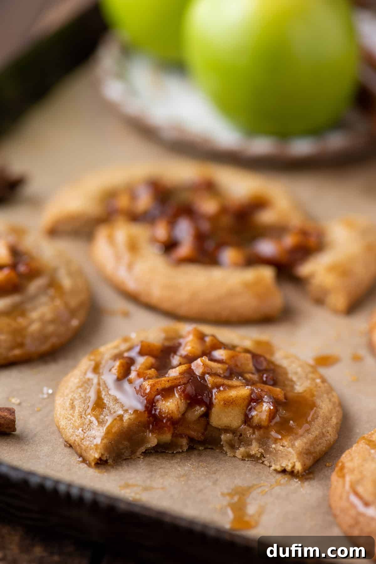 A close-up shot of a bitten apple pie cookie, revealing its soft, chewy texture, spiced apple filling, and a glistening caramel drizzle on top.