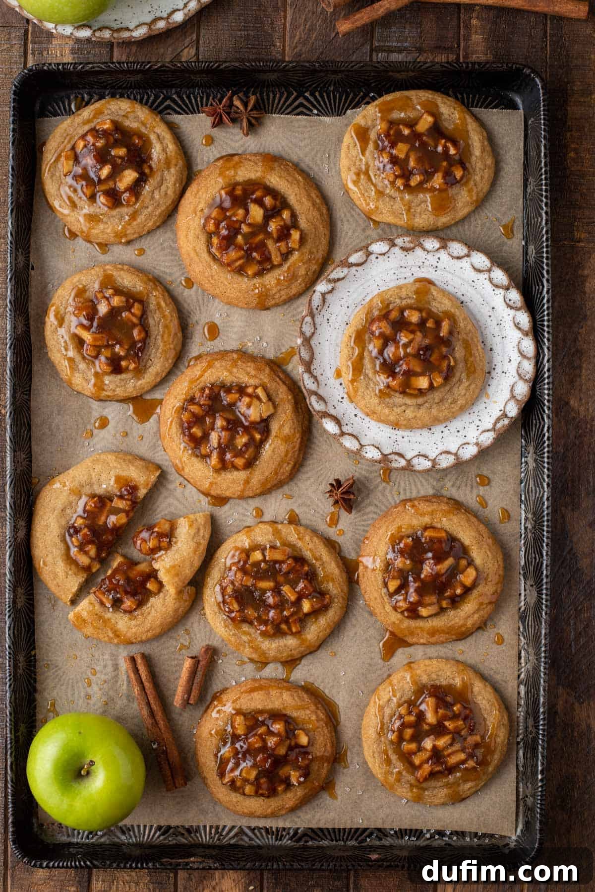 An overhead view of several apple pie cookies arranged on a parchment-lined baking sheet, with one cookie placed on a small white plate, ready to be enjoyed.