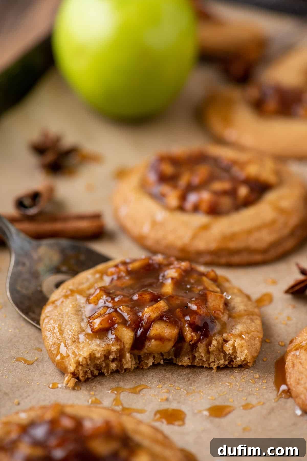 A close-up of apple pie cookies on a parchment-lined pan, with one cookie having a bite taken out of it to show the warm apple filling and soft cookie texture.