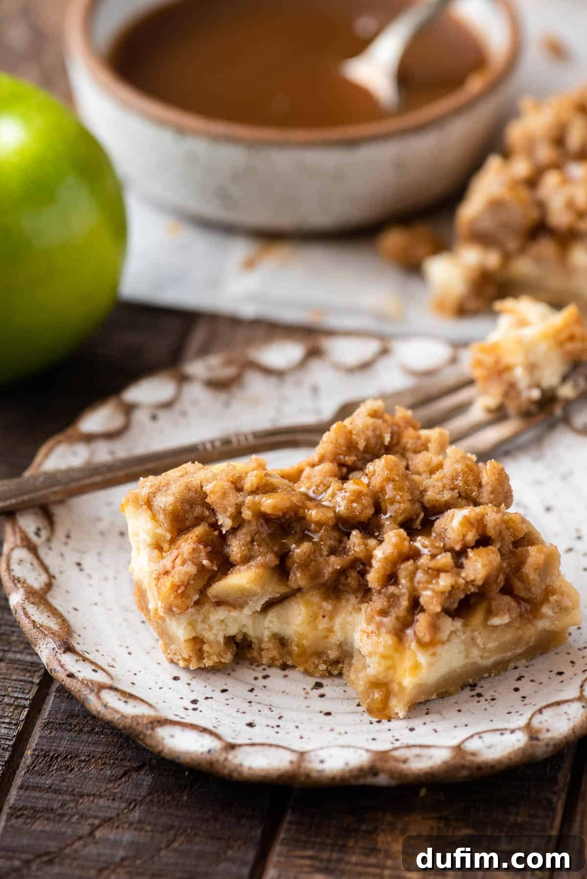 A single caramel apple cheesecake bar on a plate with a corner bite taken out, showing the layers and creamy texture.
