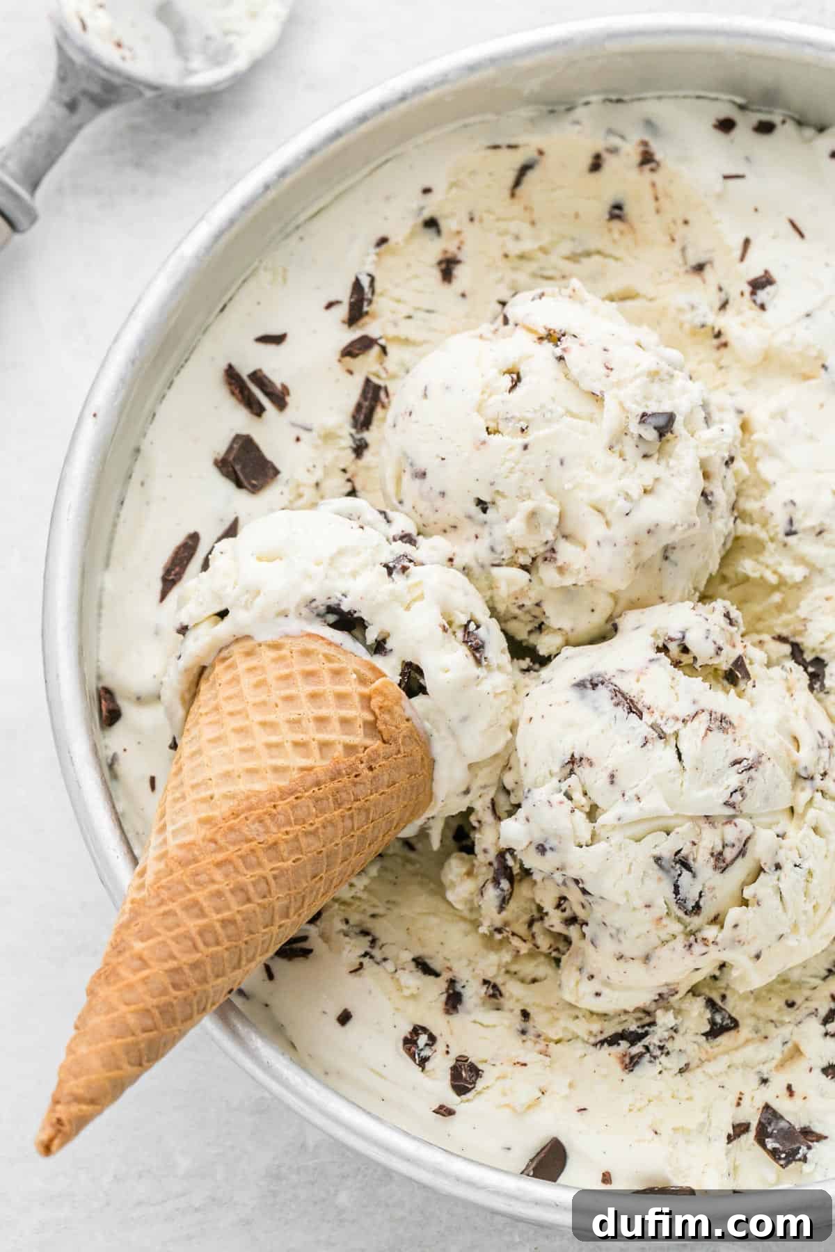 No-churn chocolate chip ice cream in a round cake pan with three scoops and one ice cream cone, garnished with chocolate shavings.