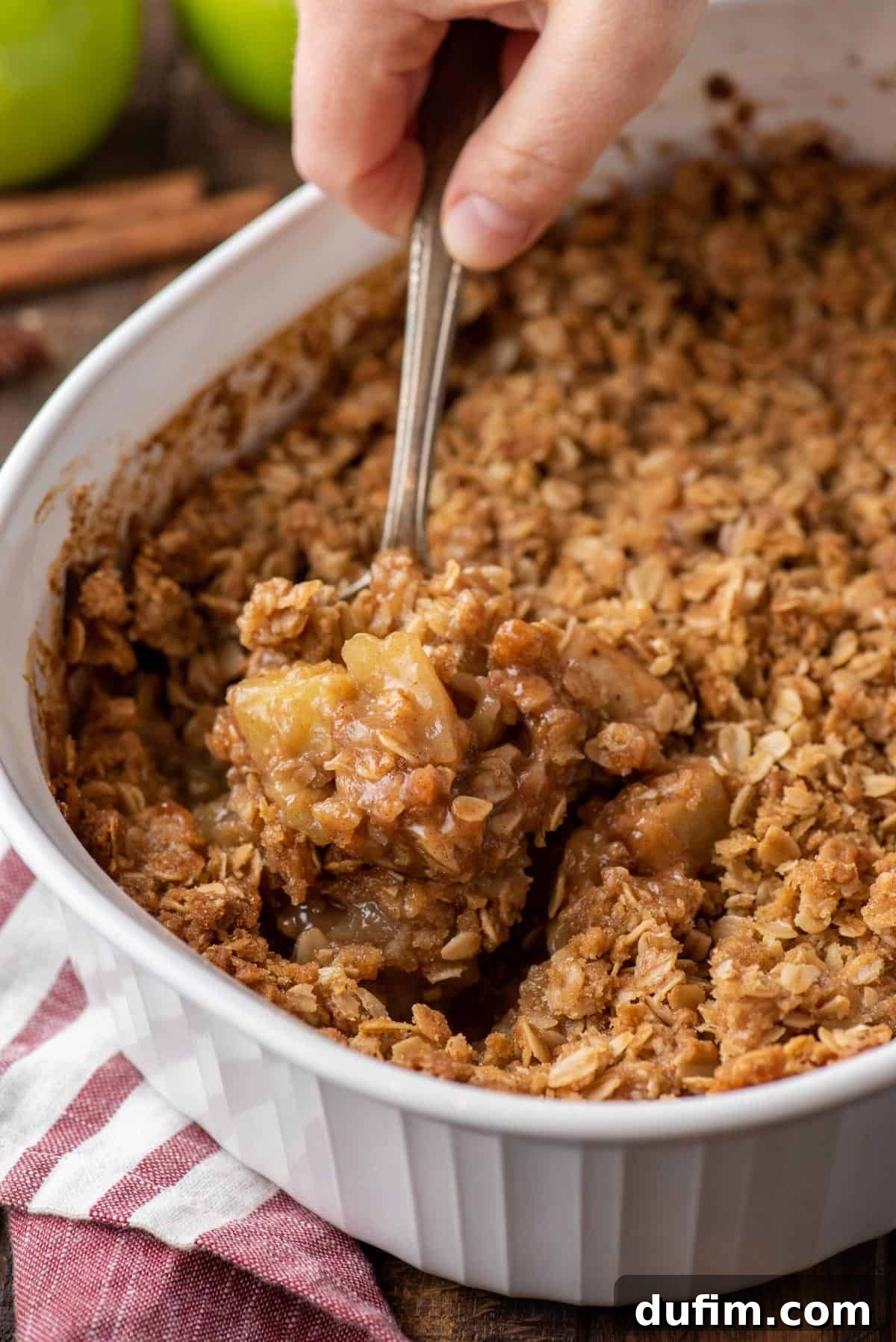 A spoon expertly scooping a generous portion of warm caramel apple crisp from a rustic baking dish, showcasing the golden-brown crispy topping and bubbling apple filling beneath.