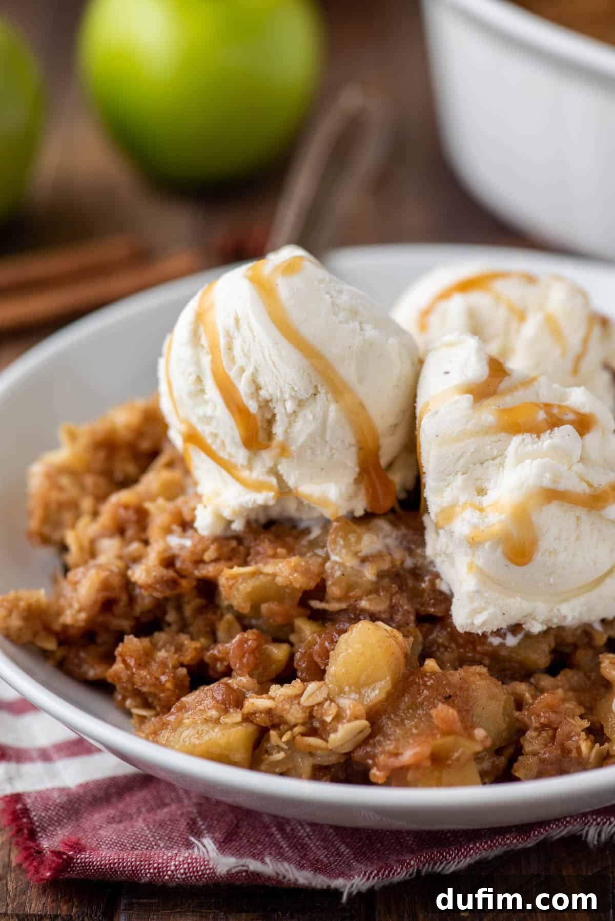 A serving of warm caramel apple crisp in a bowl, generously topped with a scoop of melting vanilla ice cream and an extra drizzle of caramel sauce, ready to be enjoyed.