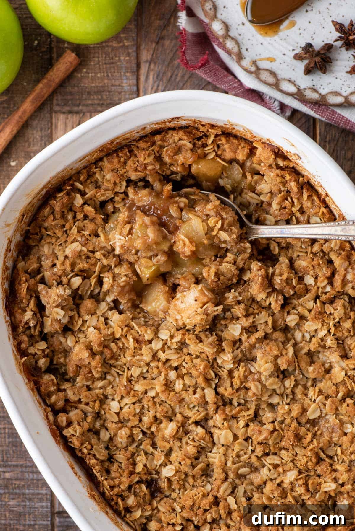 Overhead view of a golden-brown caramel apple crisp in a rectangular baking dish, with a serving spoon resting in it, revealing the rich, bubbling apple and caramel filling.