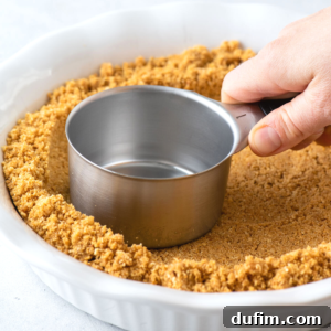 A hand presses a metal measuring cup into a pie dish filled with crushed graham crackers, forming a perfect graham cracker crust.