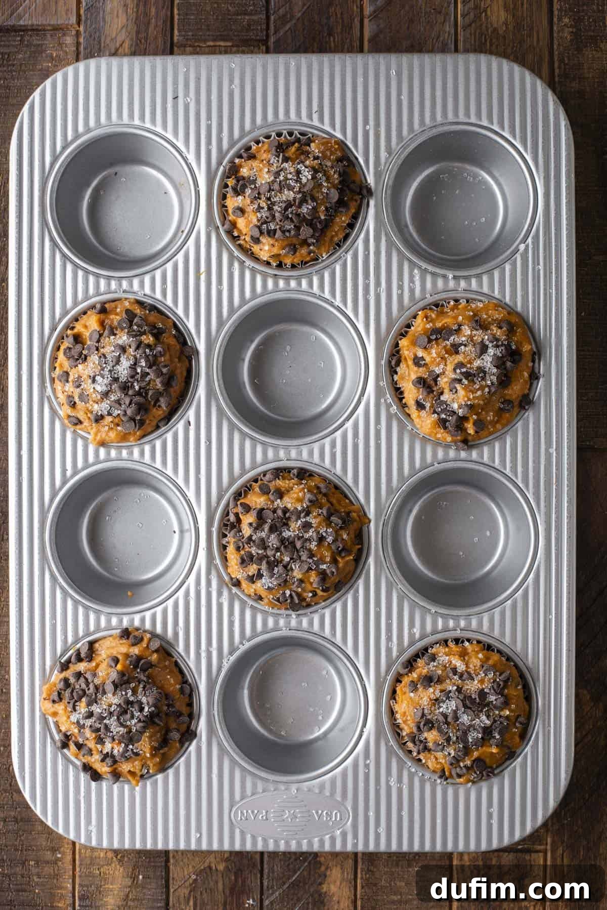 Golden Pumpkin Chocolate Chip Muffins 10 A muffin pan neatly arranged with eight filled cups of Pumpkin Chocolate Chip Muffins batter, each generously topped with chocolate chips and a delicate sprinkle of sugar. Four adjacent cups in the tin remain empty, all situated on a rustic wooden surface, showcasing strategic placement for optimal baking.
