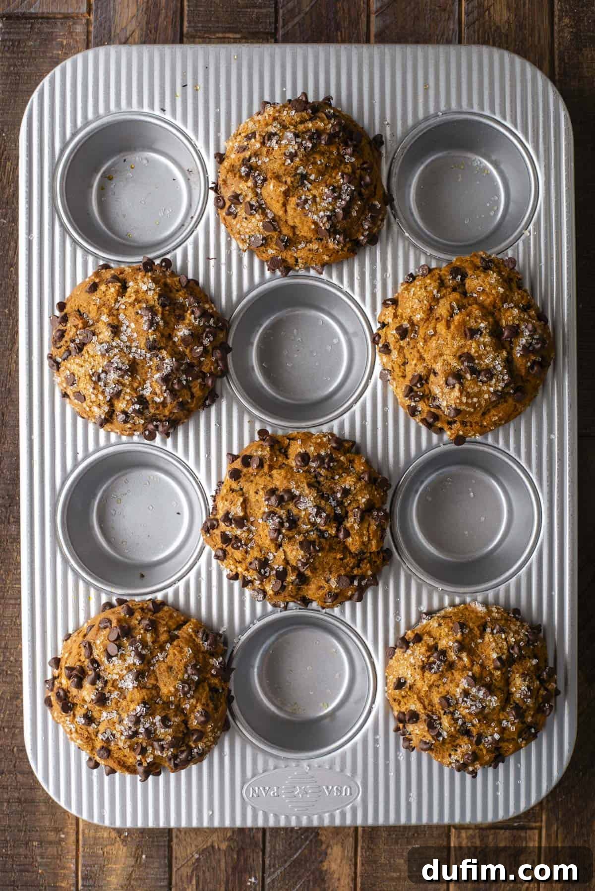 A muffin tin displays seven beautifully arranged Pumpkin Chocolate Chip Muffins, generously sprinkled with glistening sugar, positioned on a rustic wooden surface. Five other spaces in the tin are empty, suggesting a strategic baking method.