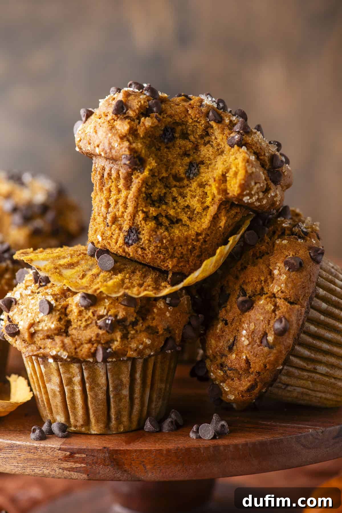 A close-up of beautifully baked Pumpkin Chocolate Chip Muffins. One muffin, positioned centrally, has a bite taken out, clearly revealing its moist, tender interior. These muffins are elegantly stacked on a rustic wooden stand, set against a warm, earthy brown background, creating an inviting and wholesome visual.