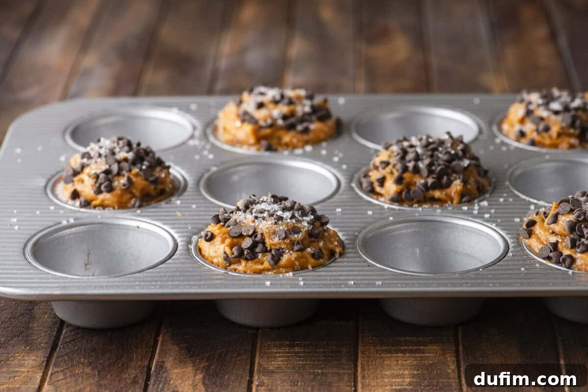 A metal muffin tin holds six unbaked Pumpkin Chocolate Chip Muffins batter mounds topped with chocolate chips and sprinkled with coarse sugar, resting on a rustic wooden surface. The alternate spaces in the tin are intentionally left empty.