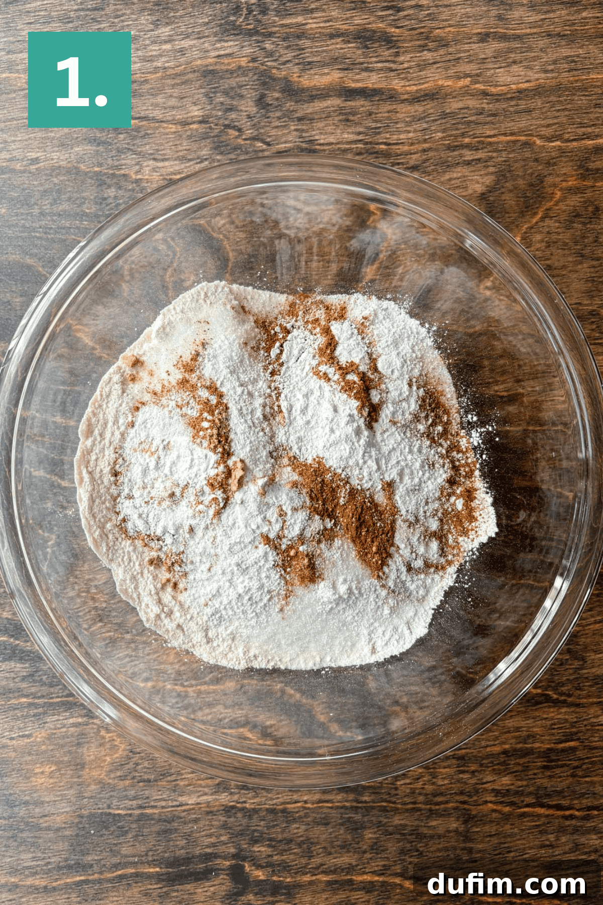 A clear glass bowl containing a mixture of flour and various spices, all key ingredients for delicious Pumpkin Chocolate Chip Muffins, sits on a warm wooden surface. The image is clearly labeled with a green box and the number 1 in the top left corner, indicating the first step in the baking process.