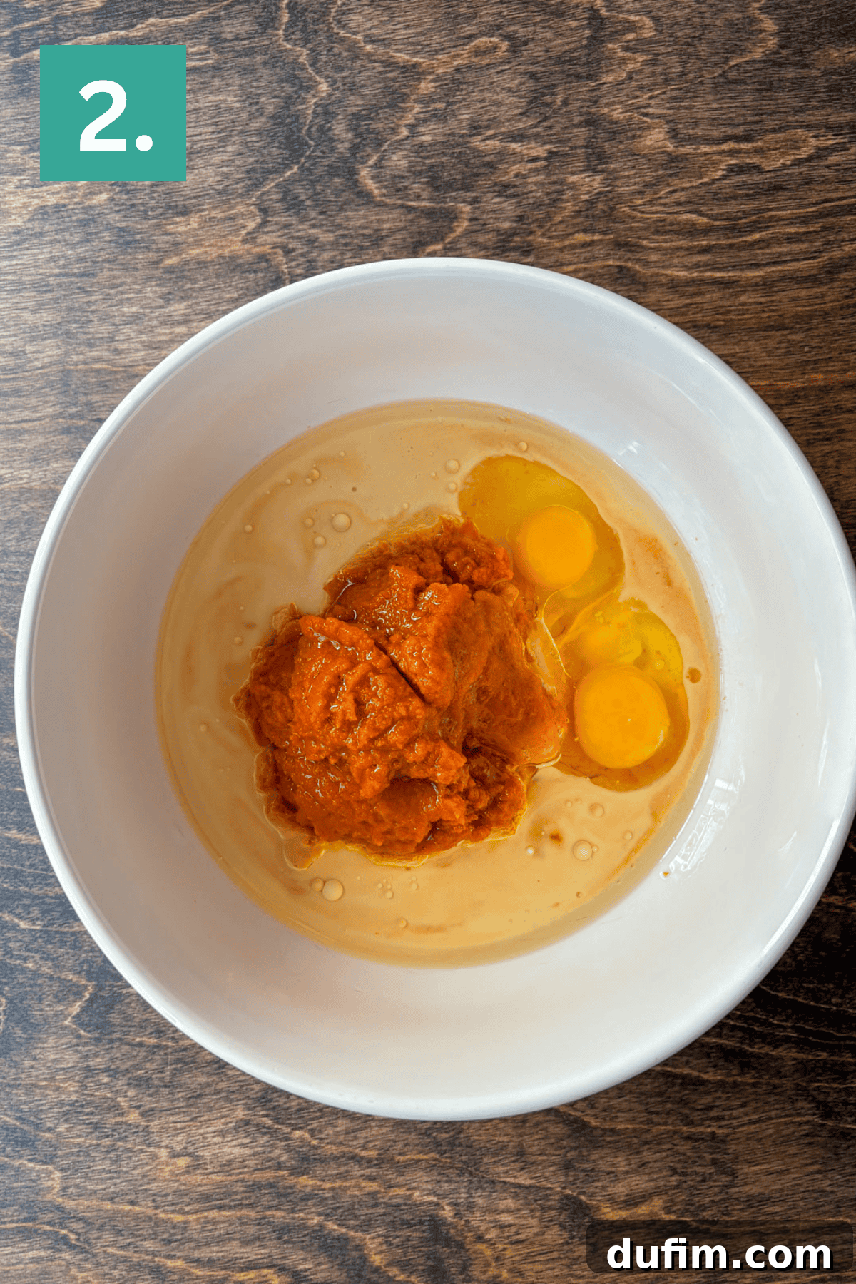 A pristine white bowl on a rustic wooden surface showcases two perfectly cracked raw eggs nestled beside a generous scoop of vibrant orange pumpkin puree, all prepared for mixing into Pumpkin Chocolate Chip Muffins. A small green box labeled with the number 2 is positioned in the top left corner, denoting the second stage of the recipe.