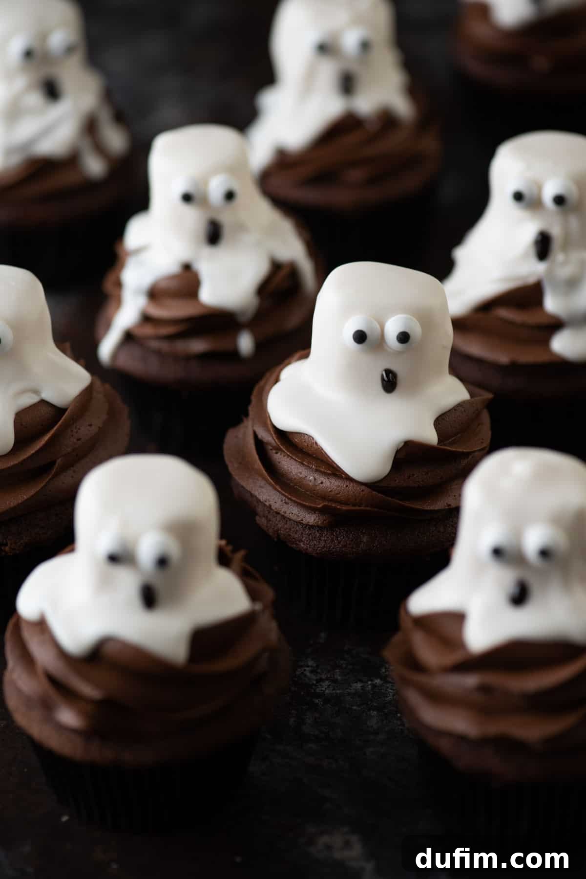 Homemade Ghost Cupcakes for Halloween Celebration An assortment of delicious ghost cupcakes, featuring rich dark chocolate bases and white marshmallow ghost decorations complete with candy eyes and drawn mouths, artfully arranged on a dark wooden background