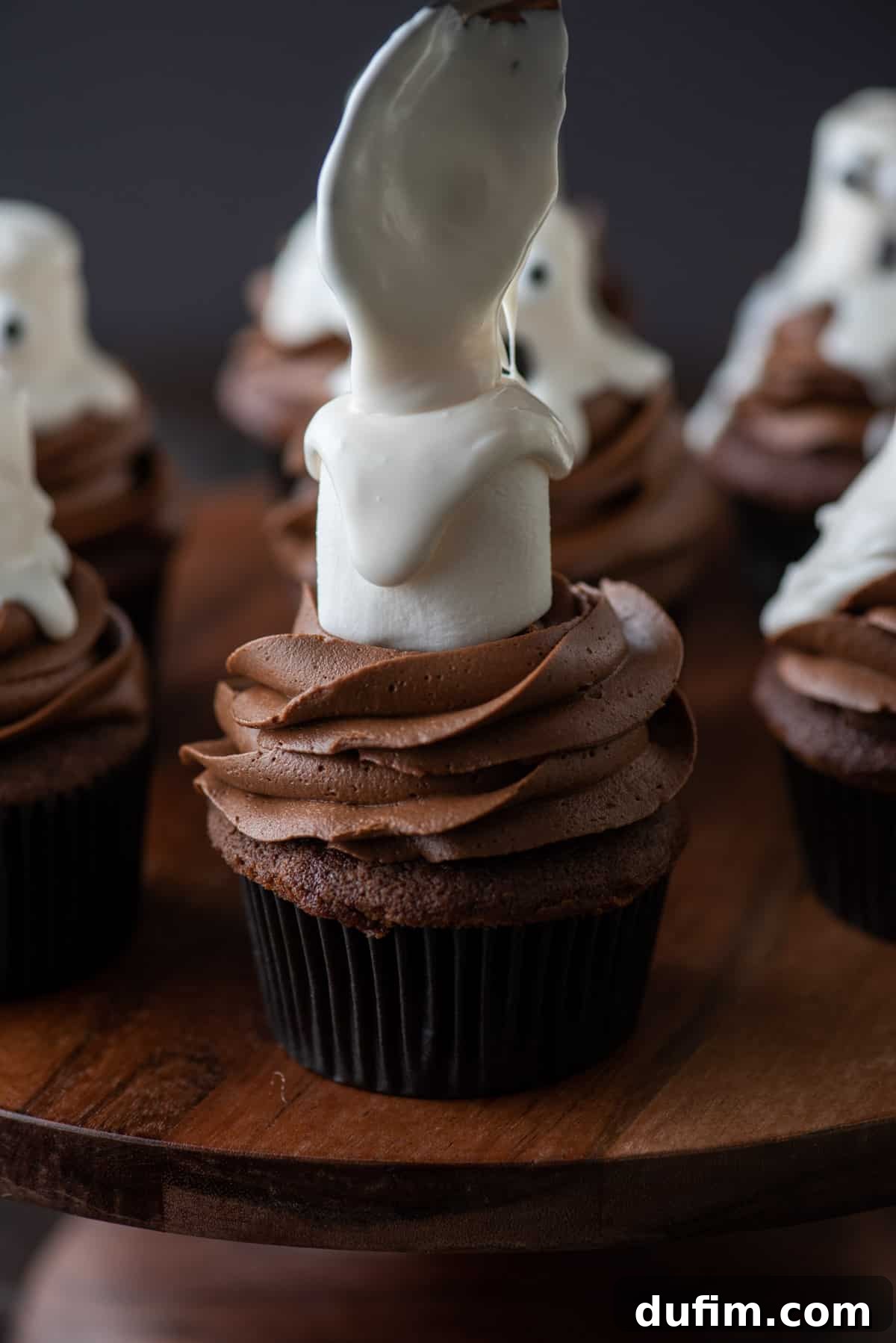 Decorating Ghost Cupcakes - Glaze Application A detailed step-by-step image showing melted white frosting being carefully drizzled over a large marshmallow that is placed on top of a chocolate cupcake, illustrating the process of forming the ghost shape