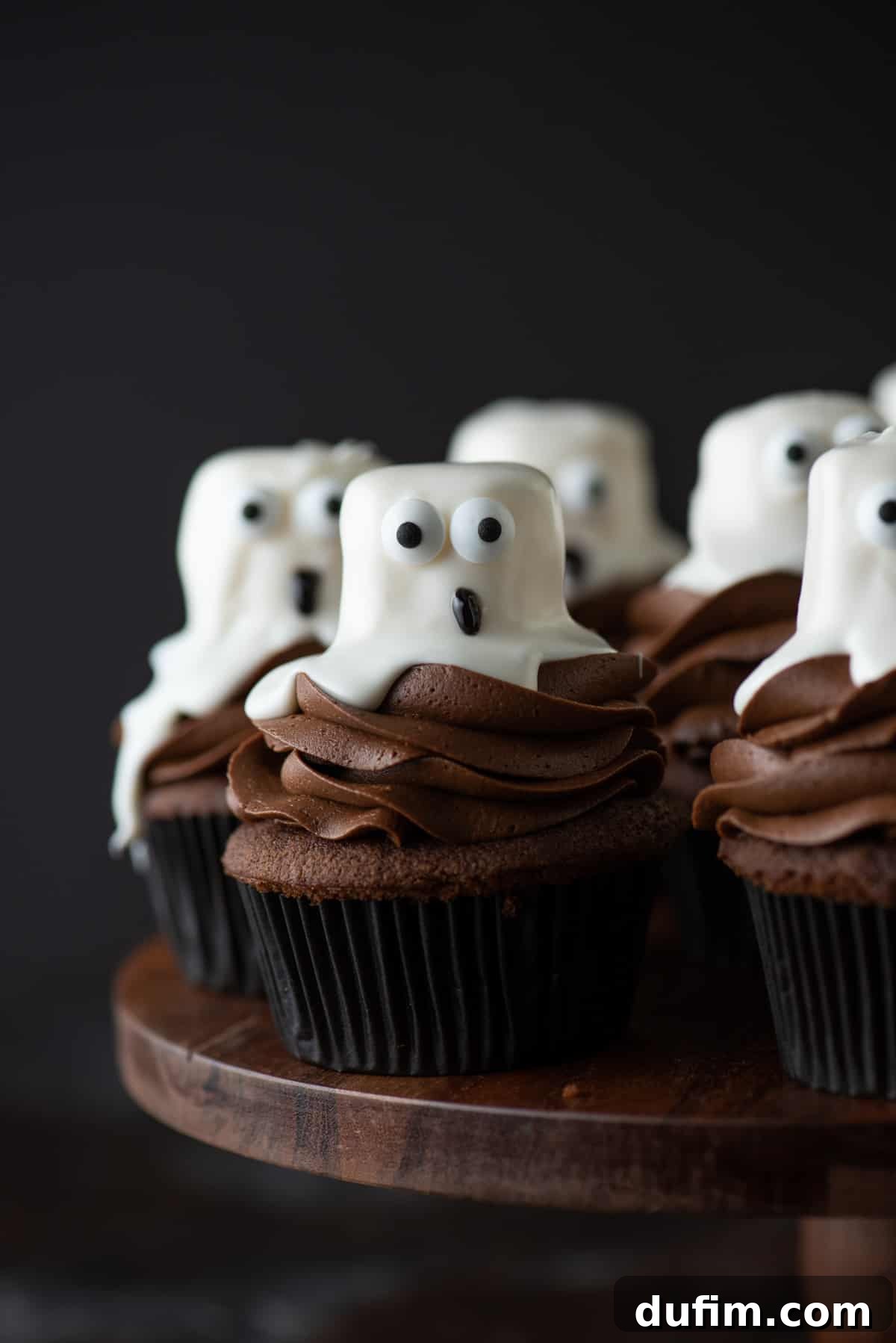 Finished Halloween Ghost Cupcakes Display A charming display of fully decorated ghost cupcakes, featuring delightful marshmallow ghosts with candy eyes and black icing mouths, elegantly arranged on a rustic wooden cake stand, perfect for any Halloween gathering