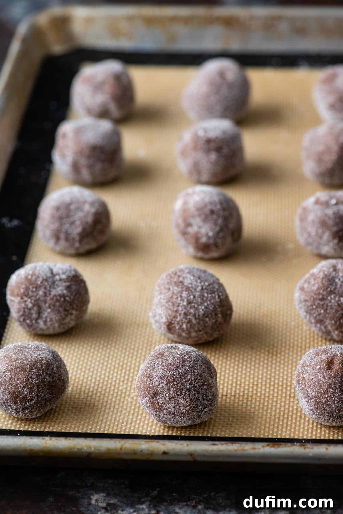 Small balls of chocolate cookie dough, perfectly rolled in granulated sugar, waiting on a baking sheet before entering the oven.