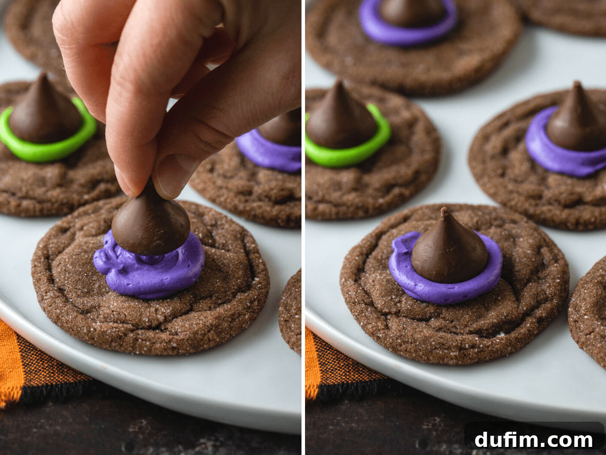 A hand gently placing a shiny Hershey's Kiss onto a dollop of vibrant purple frosting on a chocolate cookie, forming a witch's hat.