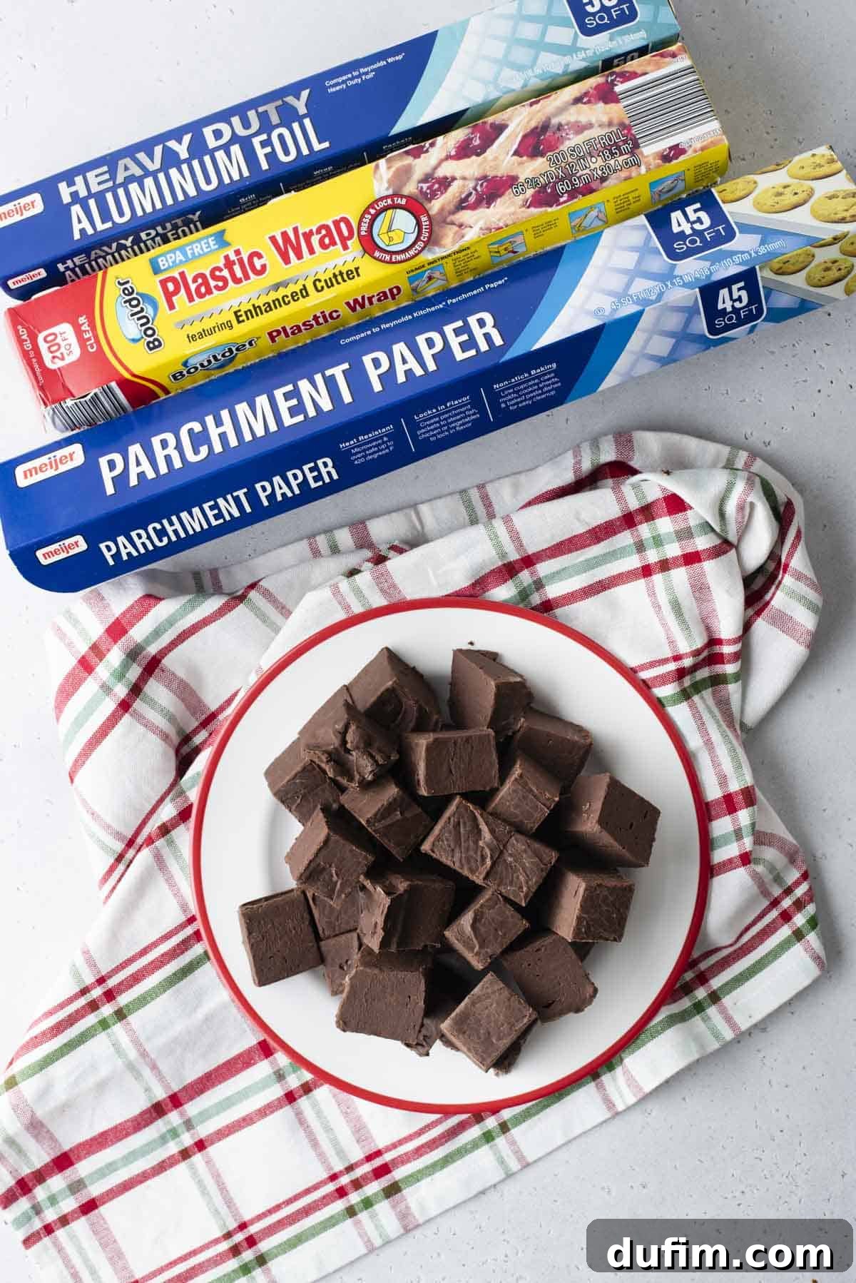 A display of essential fudge freezing tools: aluminum foil, plastic wrap, parchment paper boxes, and a plate of freshly cut chocolate fudge.