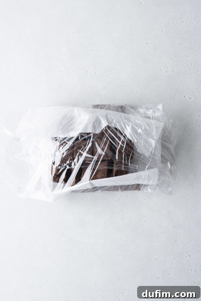Several layers of chocolate fudge, separated by parchment paper, are being carefully wrapped in plastic wrap for optimal freezing.