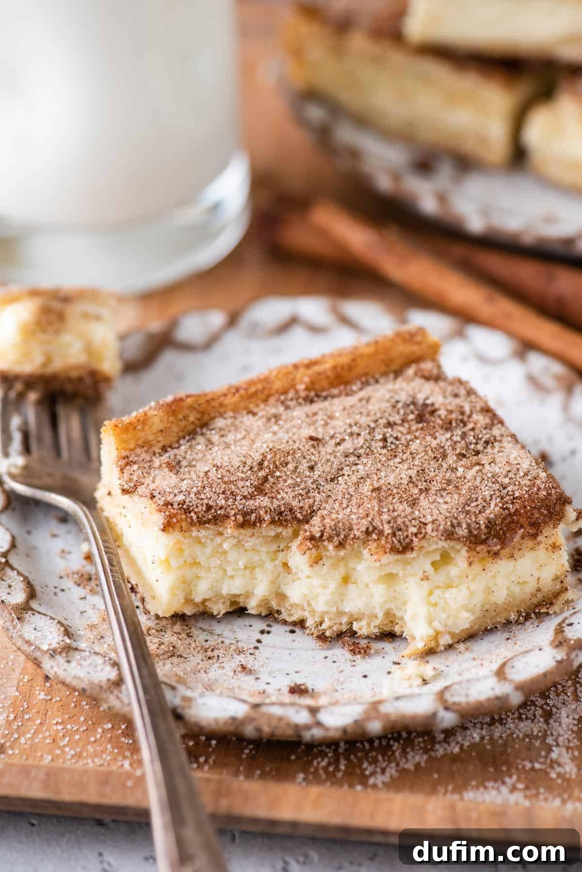 With a buttery, flaky crust, cinnamon sugar topping, and tangy cheesecake filling, this Churro Cheesecake is easy to make & ridiculously good. A churro cheesecake bar on a plate with a bite taken out, showing the creamy interior and flaky crust