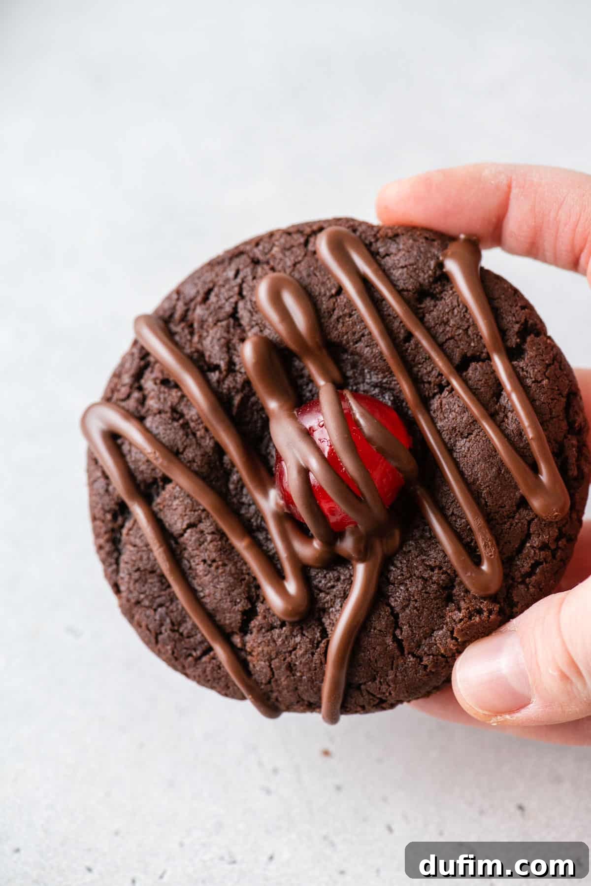 A hand holding a perfectly baked chocolate cherry cookie, showcasing the rich chocolate and red cherry center.