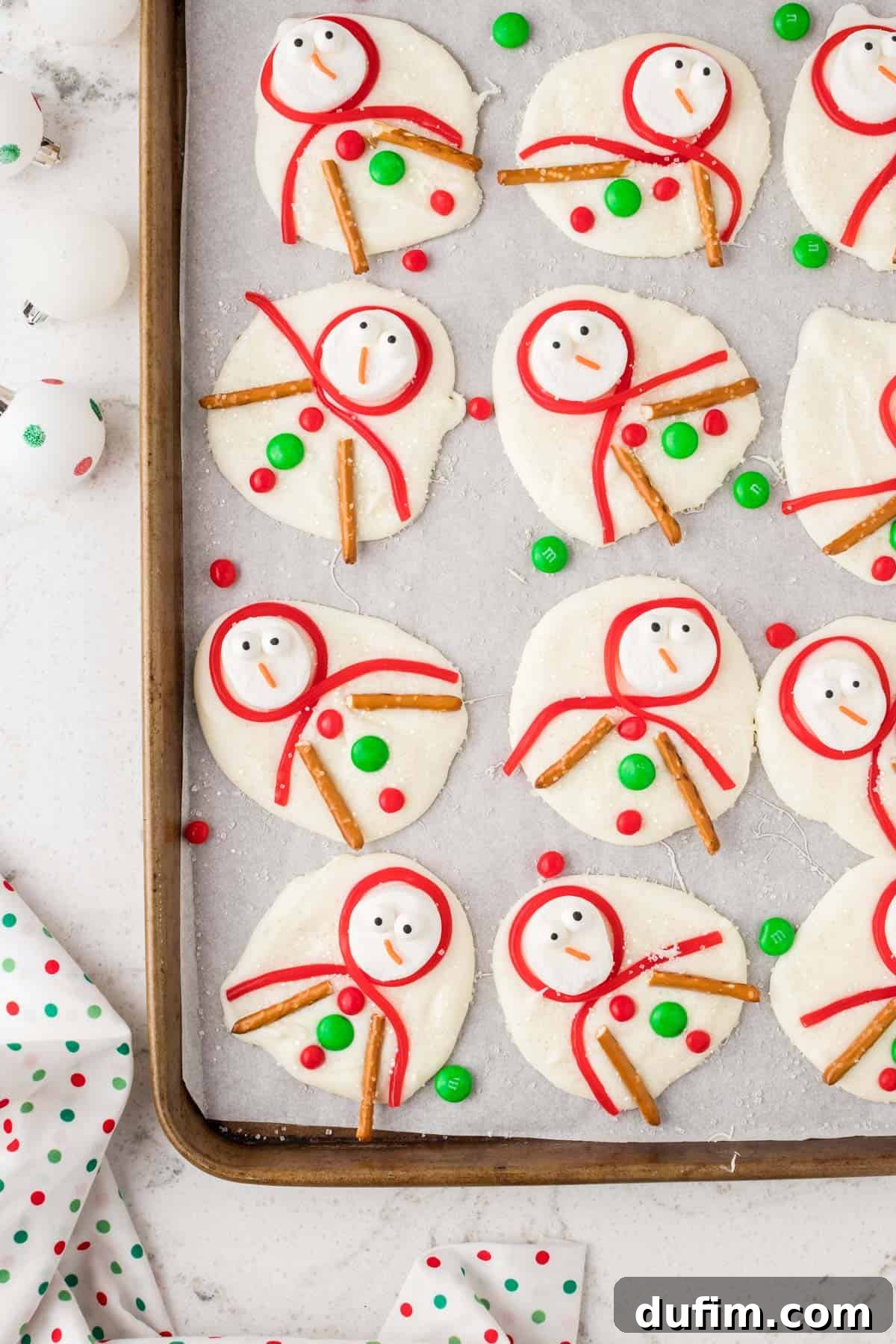 Melted Snowman Bark artfully arranged on a pristine white baking sheet lined with parchment paper, showcasing charming marshmallow snowmen amidst a sprinkle of sugary 'snow'.