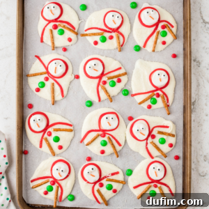 A baking sheet with twelve whimsical melted snowman bark pieces, each featuring a marshmallow head with candy eyes and an orange jimmy nose, a red Twizzler scarf, pretzel stick arms, and colorful candy buttons. Red and green M&Ms are scattered around, enhancing the festive look.