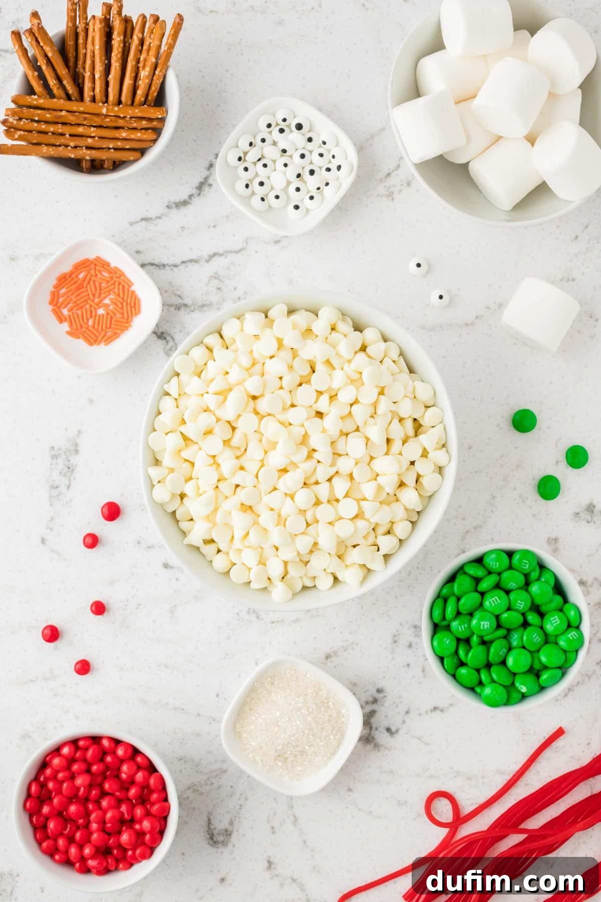 A collection of ingredients neatly arranged in white bowls on a white background, including marshmallows, Twizzlers, candy eyeballs, orange jimmies, white chocolate chips, cinnamon hots, green M&M's, pretzel sticks, and sanding sugar.